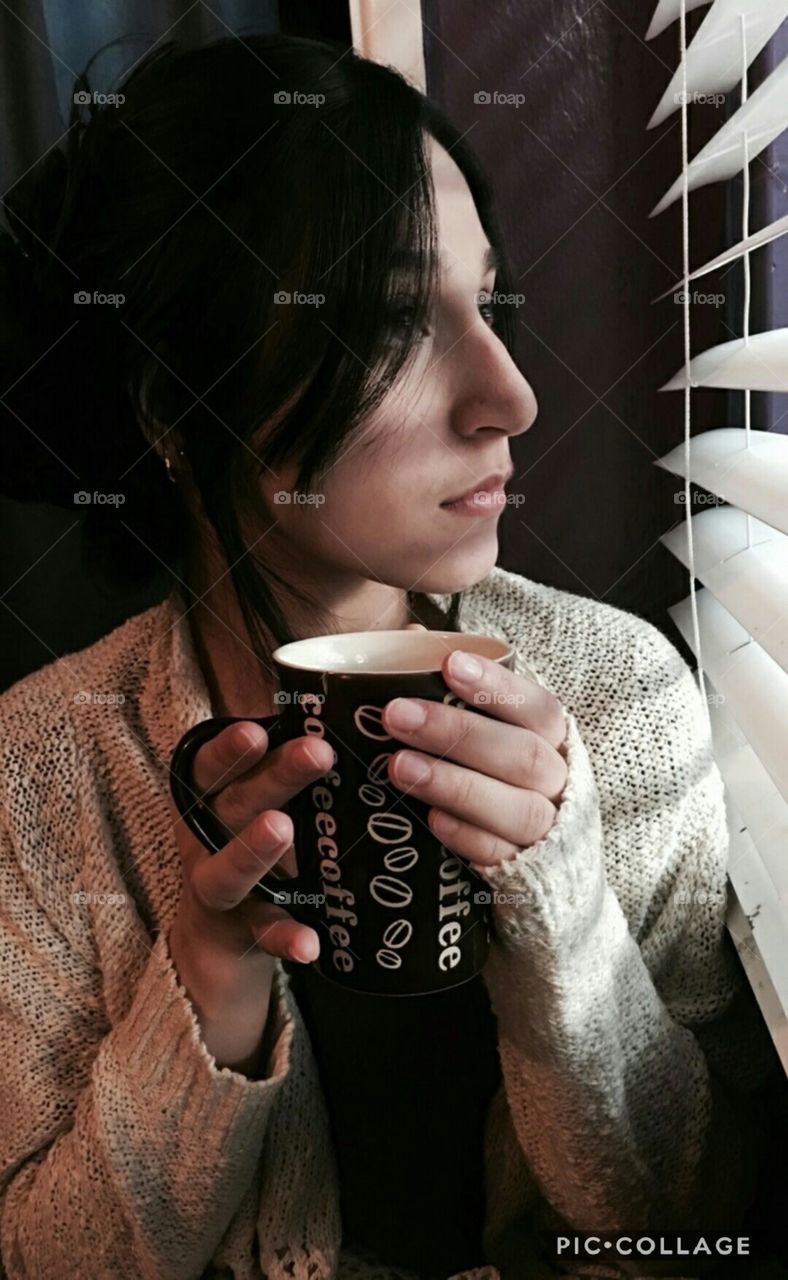 pearning through the window at the dark grey skies above, Drinking a cup of hot coffee to keep her warm