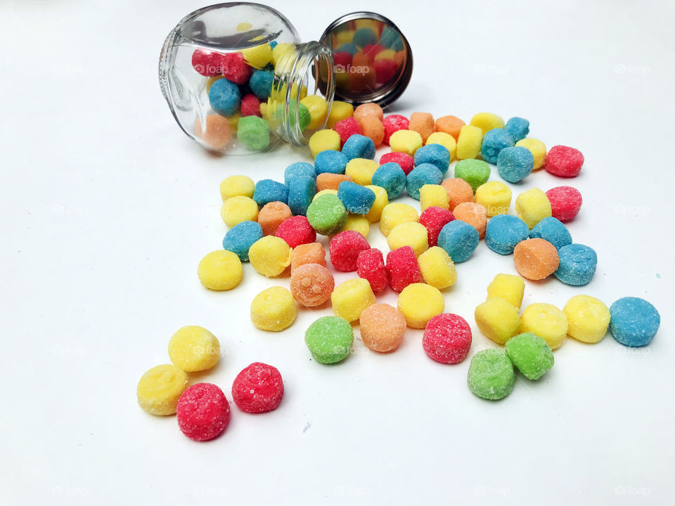 Sour chewy gummies in green, turquoise, red, yellow & orange flowing out of a small overturned glass jar. The up turned lid reflects the candies. The candies have little smily faces & an orange one in the foreground smiles at the viewer! 😋