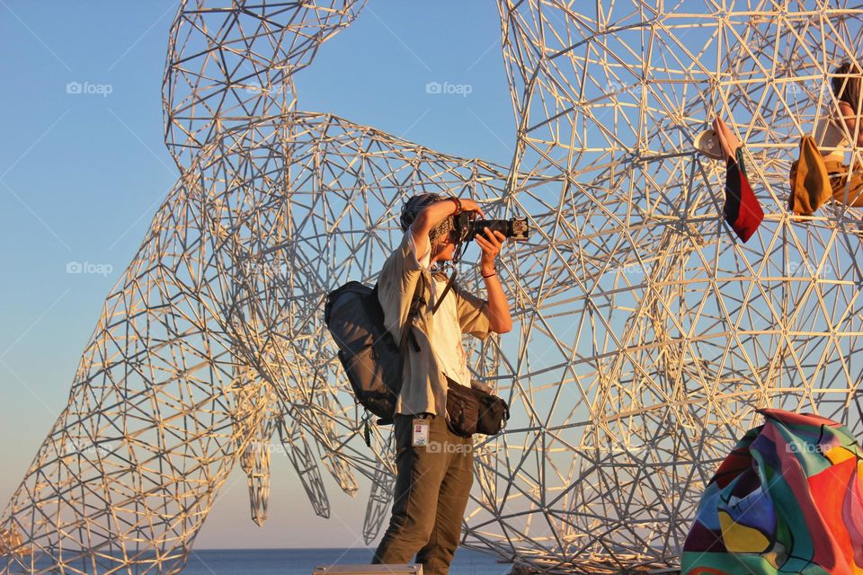 a photographer against the backdrop of a strange metal structure near the sea takes a photo at sunset