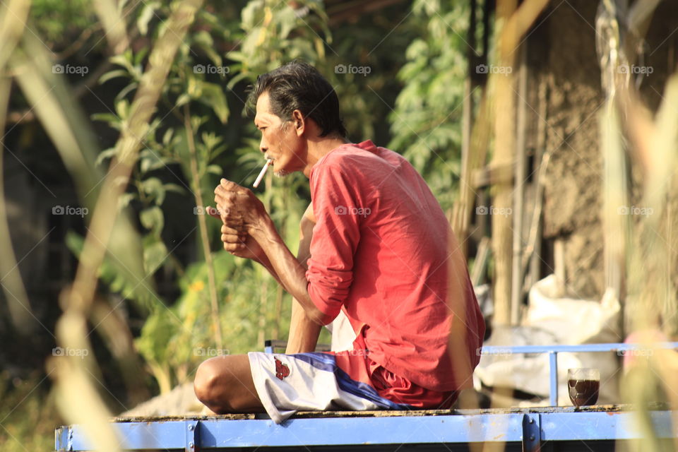 An old man wearing a red shirt sits relaxing while lighting a cigarette