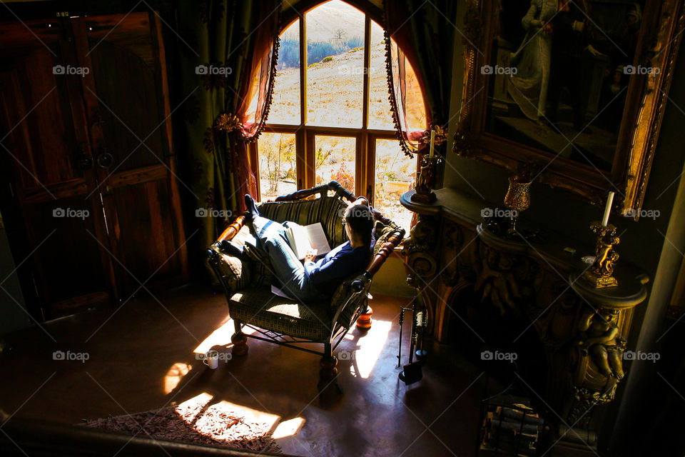 One of my favourite hobbies is reading with a cup of coffee. Image of a woman by a large window with light streaming in from outside, reading a book in am armchair