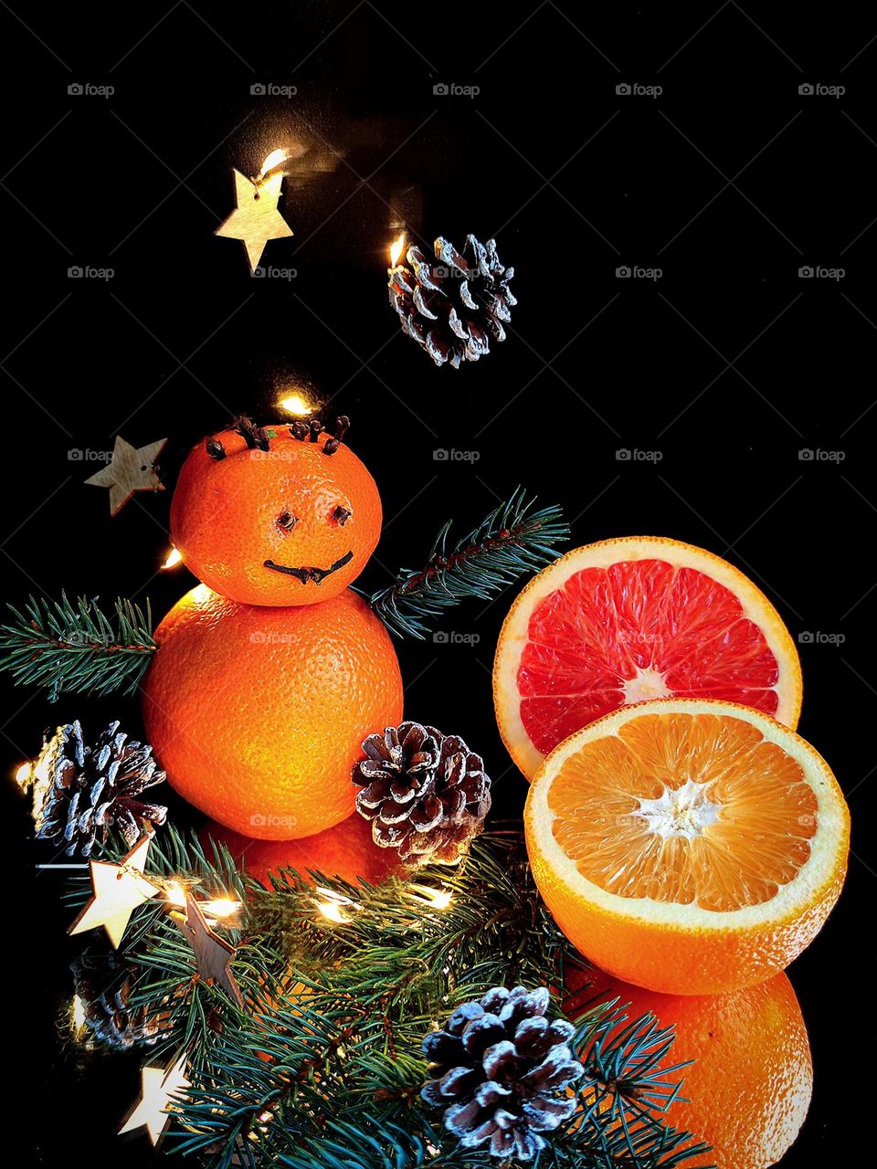 New Year card. Smiling mandarin and orange snowman on a black background and on a mirror. Spruce hands of a snowman are open to hugs. Nearby lie halves of oranges and a spruce branch. Around a luminous garland of cones.