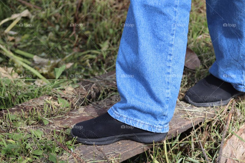 Human leg with blue jeans and black shoe in nature. Rustic plank in a green bushes 