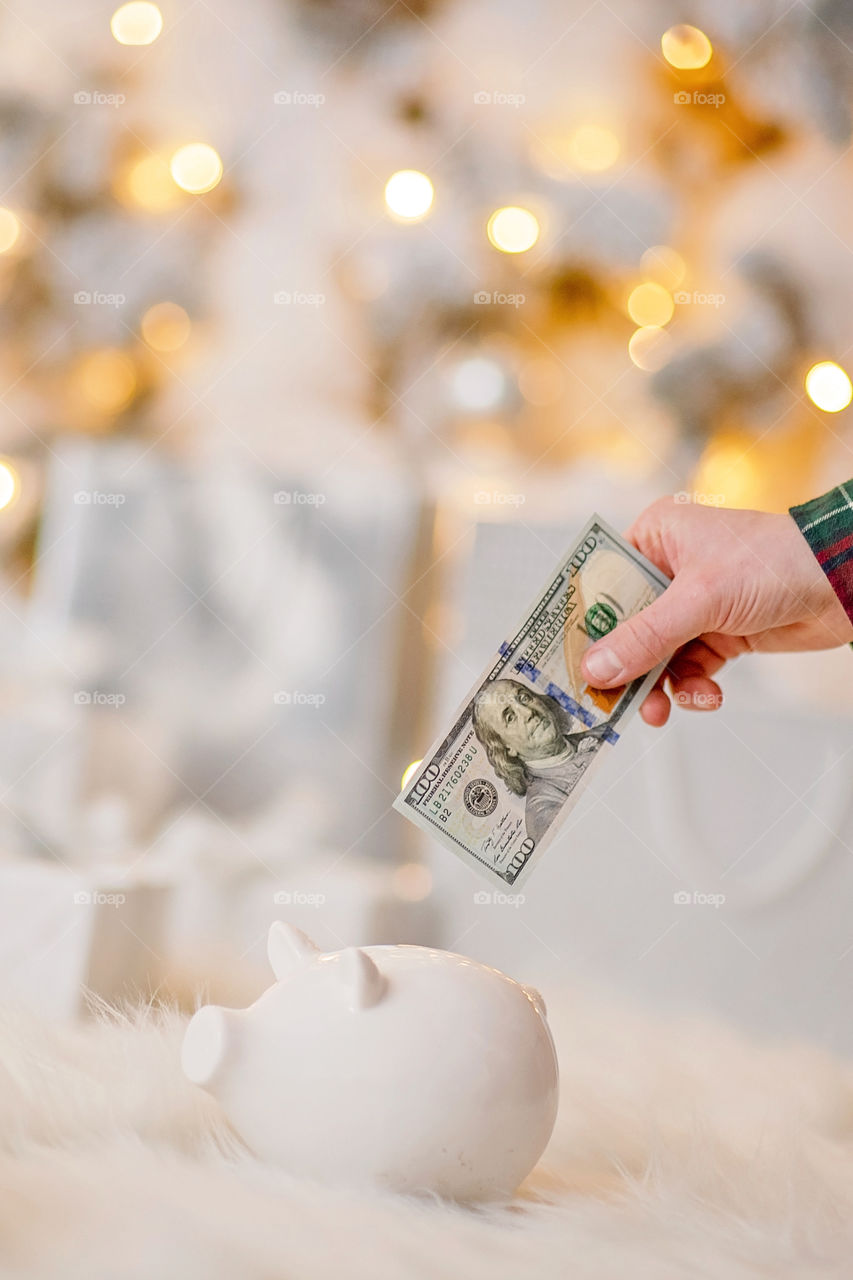 piggy bank with 100 dollars banknote in festive New Year atmosphere of scenery.  Magical bokeh with Christmas tree and bright lights.