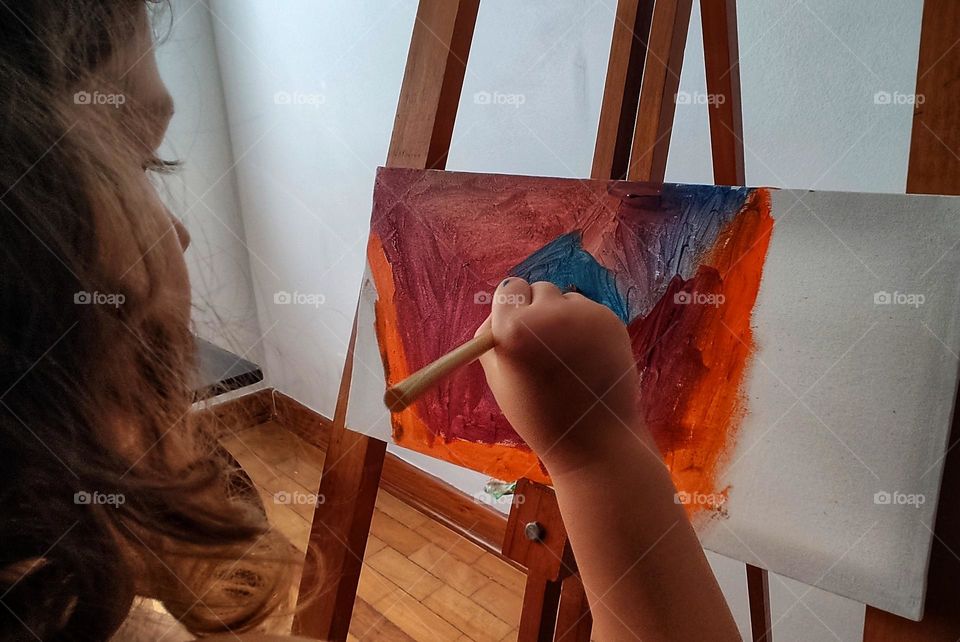 A child's hand with a brush on the background of the canvas, draws a picture in red and blue tones.