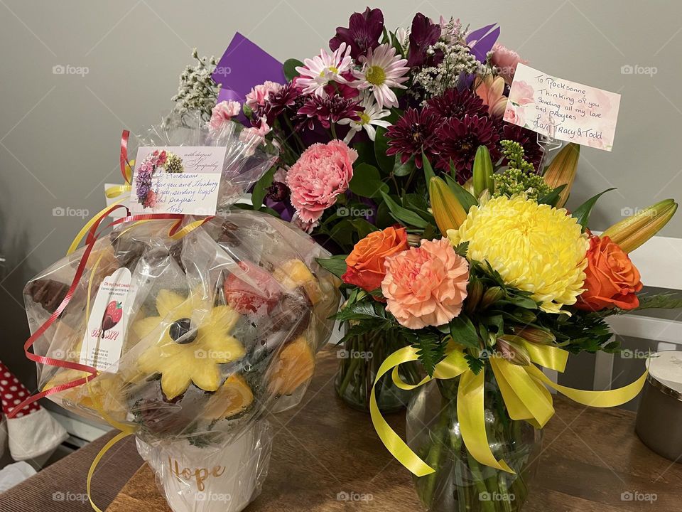 Bouquets of flowers in a variety of glass vases, one is a fruit bouquet still covered with plastic, condolence flowers, taken with my iPhone 12