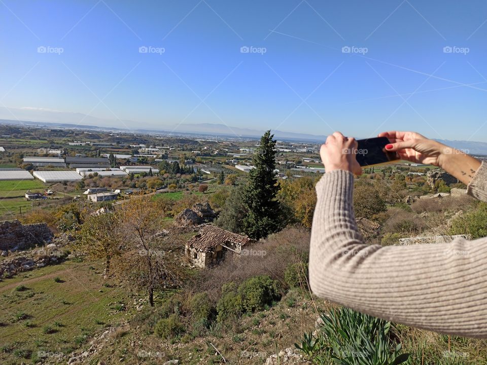 A photo of woman making a photo of landscape view of trees, fields, greenhouses and mountains from the hill