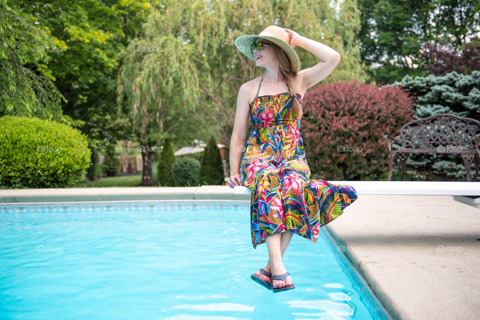 Young millennial woman wearing a dress and flip flop sandals while sitting on a diving board of a swimming pool in the summer