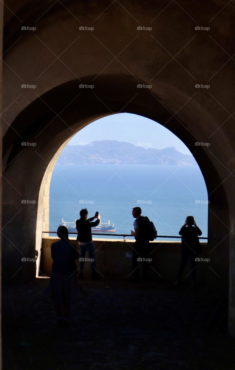 People taking photos on a big arch window viewing sea and mountains 