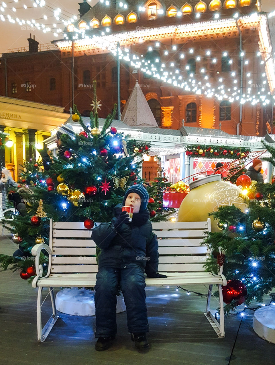 Moscow. Russia. Christmas holidays. A boy sits on a bench with a glass of hot tea at a beautifully decorated garland fair on Red Square in the evening