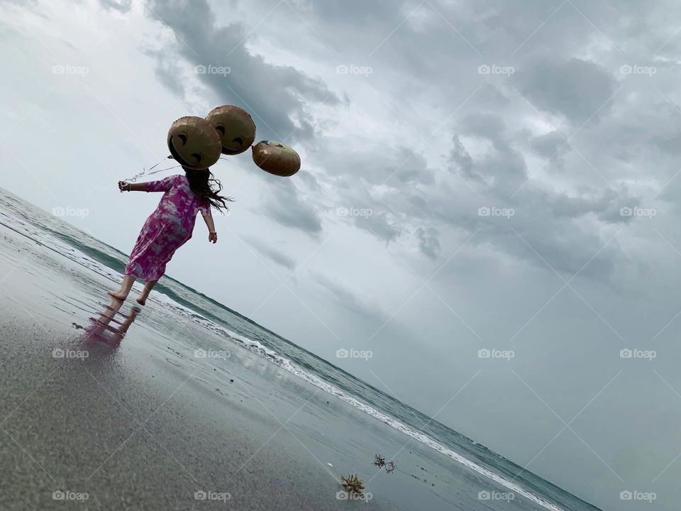 Little Girl Jumping And Running Having Fun With Helium Emoji’s Balloons On The Beach By The Ocean.