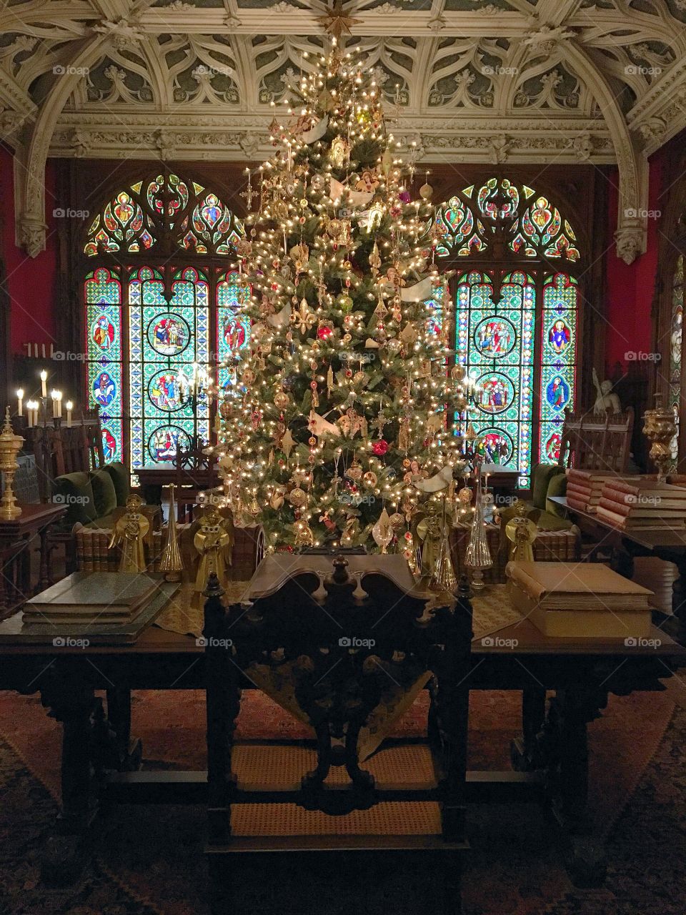 An elegantly decorated Christmas tree sits in front of an old writing desk and is illuminated by stained glass windows