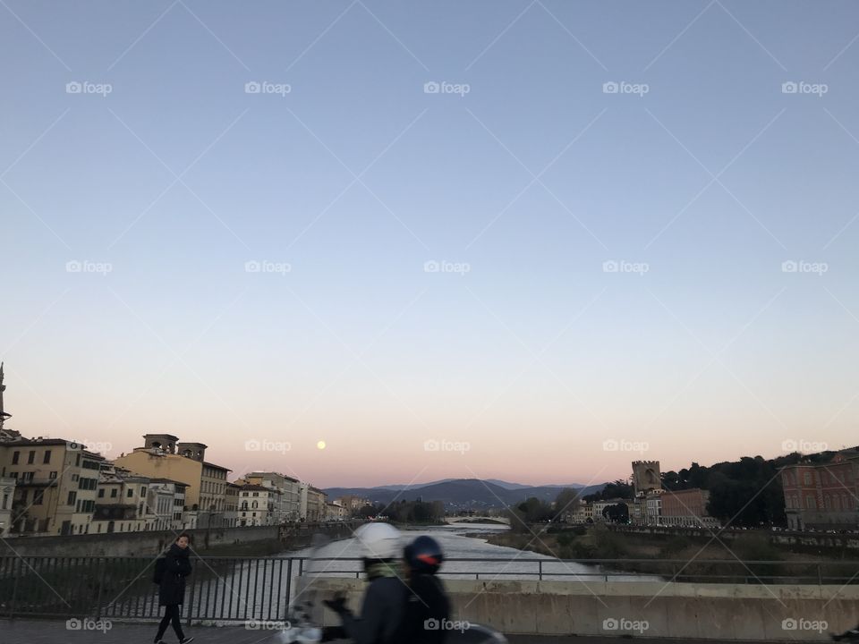 Couple driving across Ponte Alle Grazie on a scooter overlooking sunset and full moon sky in Florence, Italy,