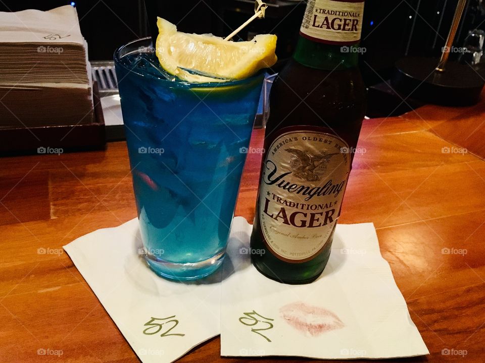 Yuenling in the bottle and a blue Long Island ice tea on bar with napkins and a kiss mark 