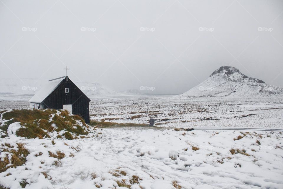 Black church in Iceland next to snow-covered hill with some grass looking out of the snow.