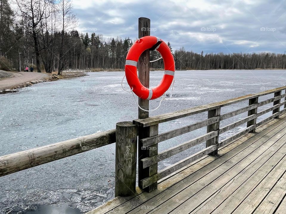 Side angle view to the bright orange red lifebuoy hanging on the pole of the wooden pier over the frozen pond water 