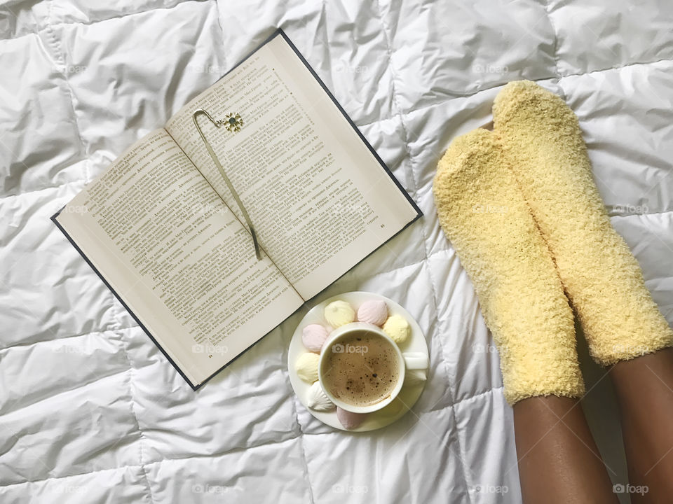 Young woman wearing cozy yellow socks enjoying reading book and coffee with candies in cozy bed in winter 