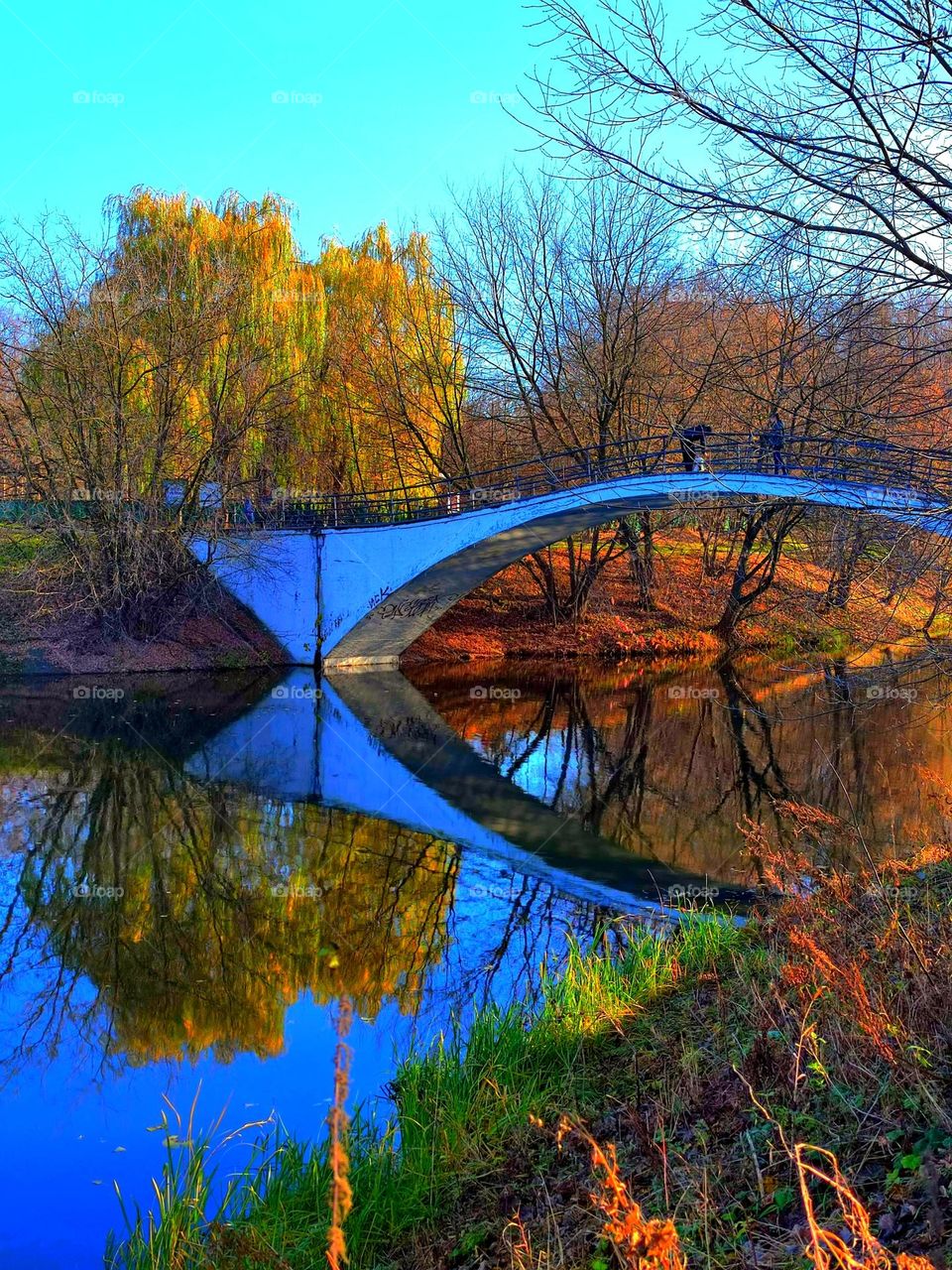 Autumn landscape.  There is a stone bridge across the river, which is reflected in the blue clear water.  On the shore there are autumn trees with yellow and red leaves, which are reflected in the blue water.