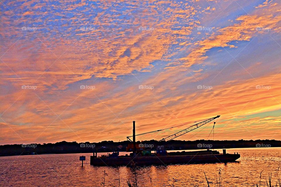 The sky displays a striking contrast of deep blues and fiery oranges, reflecting the warm hues of the setting sun on the water's surface. The silhouette of a barge is in the foreground 