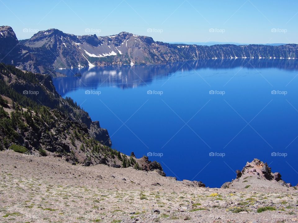 The jagged rim reflecting into the rich blue waters of Crater Lake in Southern Oregon on a beautiful summer morning with perfect clear blue skies. 