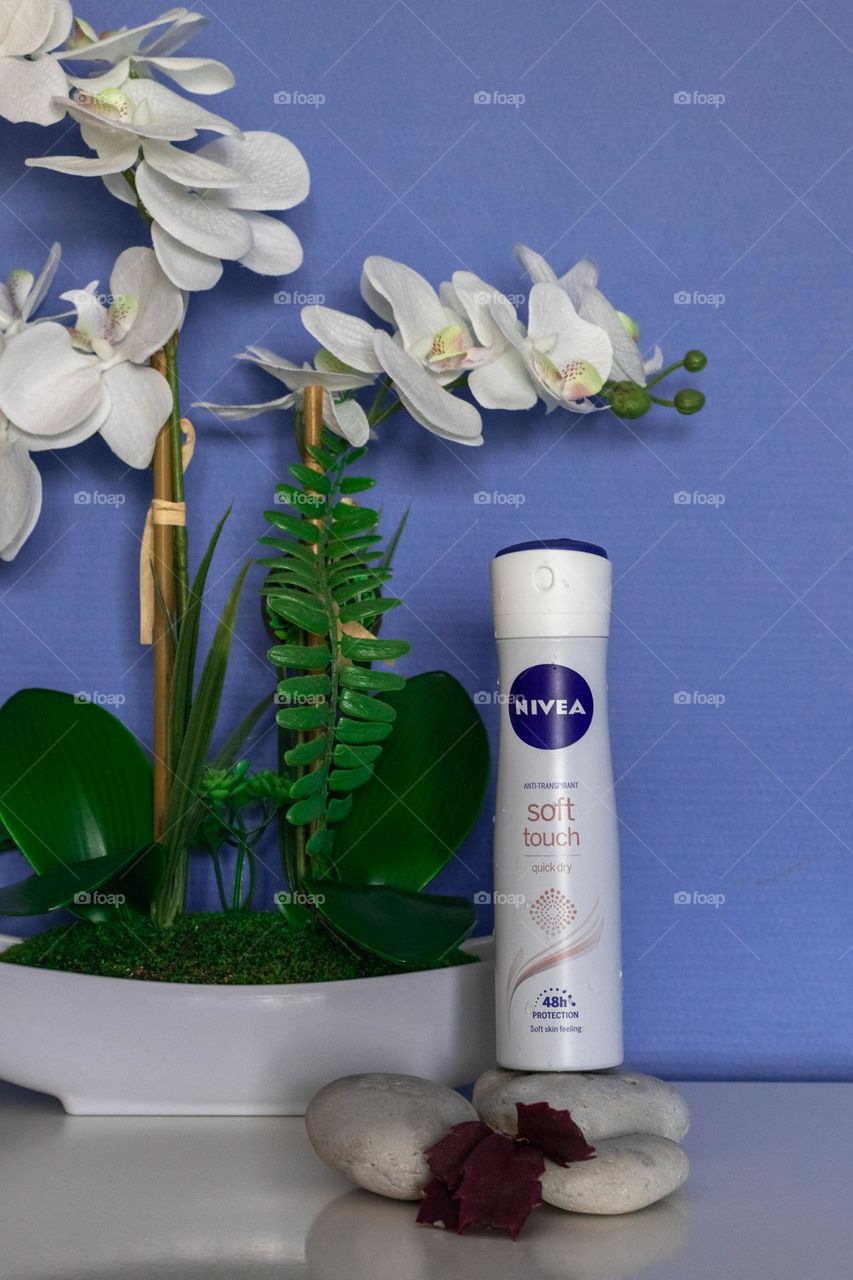 One bottle of Nivea anti-transpirant deodorant stands on white stones next to a potted orchid on a white chest of drawers against a lilac-blue wall in a bedroom, close-up side view.