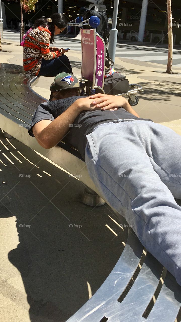 Man wearing jogging pants laying down on bench in the sun with baseball cap over face to shield the light