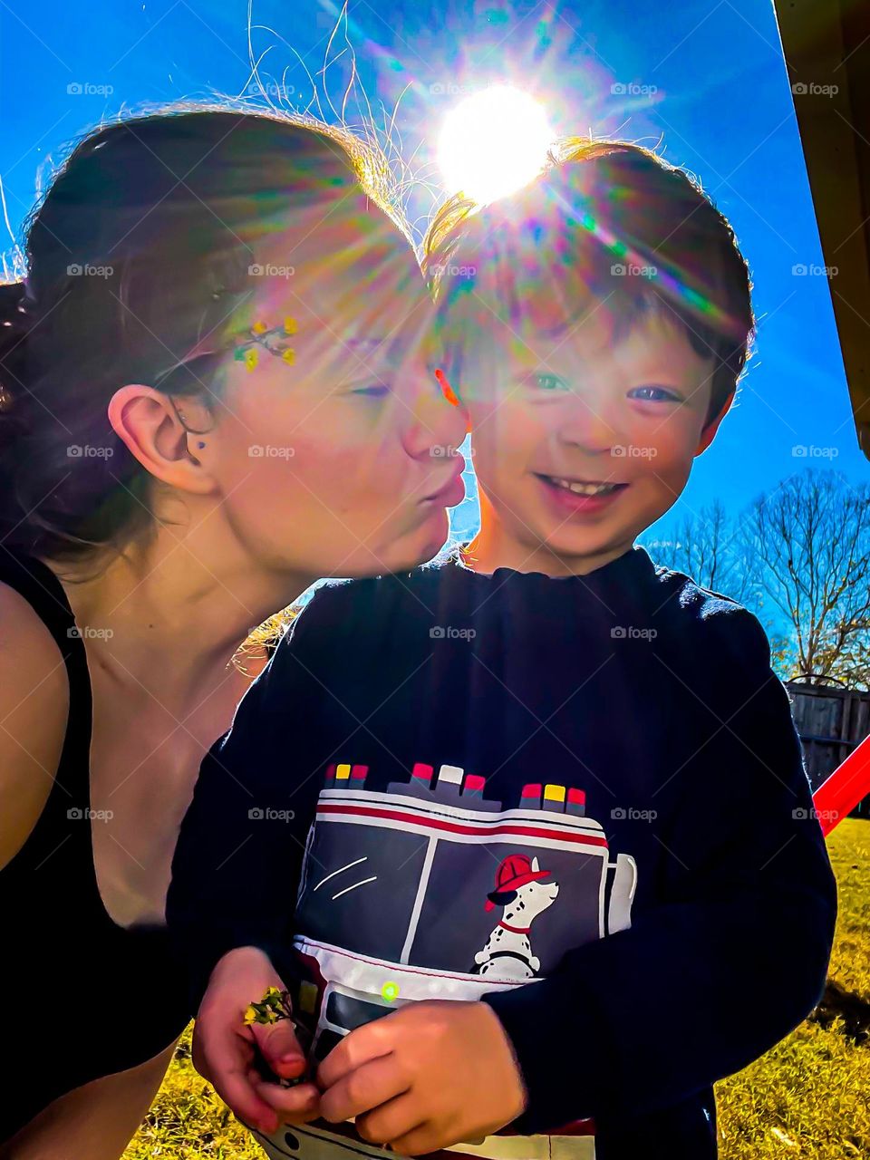 The sun shines brightly behind a mother and son. The mother, with a flower in her hair, attempts to kiss her giggling son in a show of love and comfort in nature