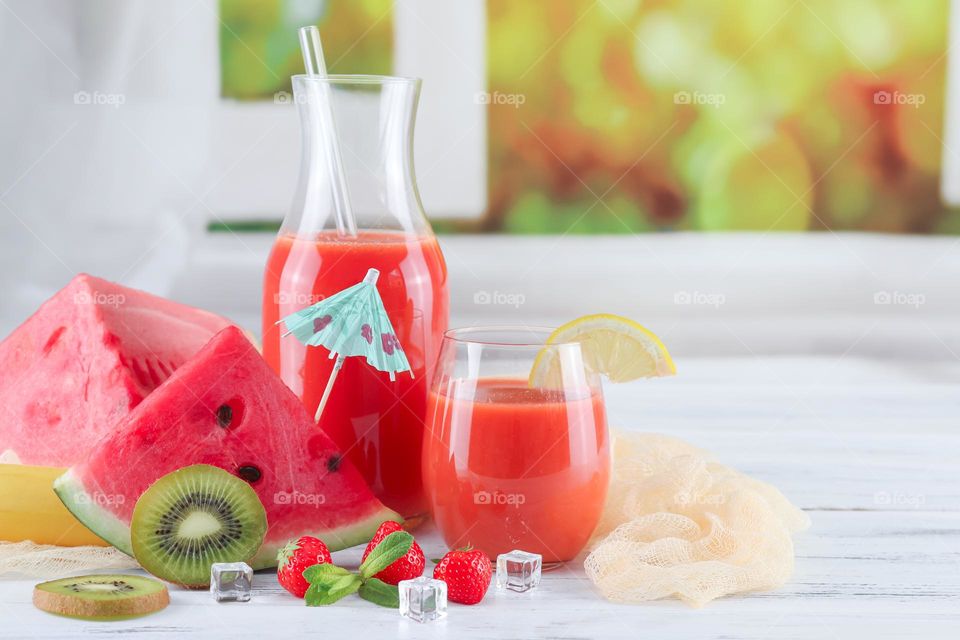 Multivitamin drink in a decanter and a glass with fresh fruits, berries: slices of kiwi, lemon and watermelon, mint leaves, rosemary and ice cubes on a light shabby table against a blurred window with a flowering field, close-up side view. Concept su