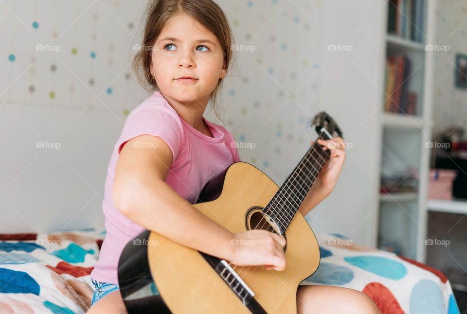 Cute tween girl in pink t-shirt play guitar sit on bed in bright room at home