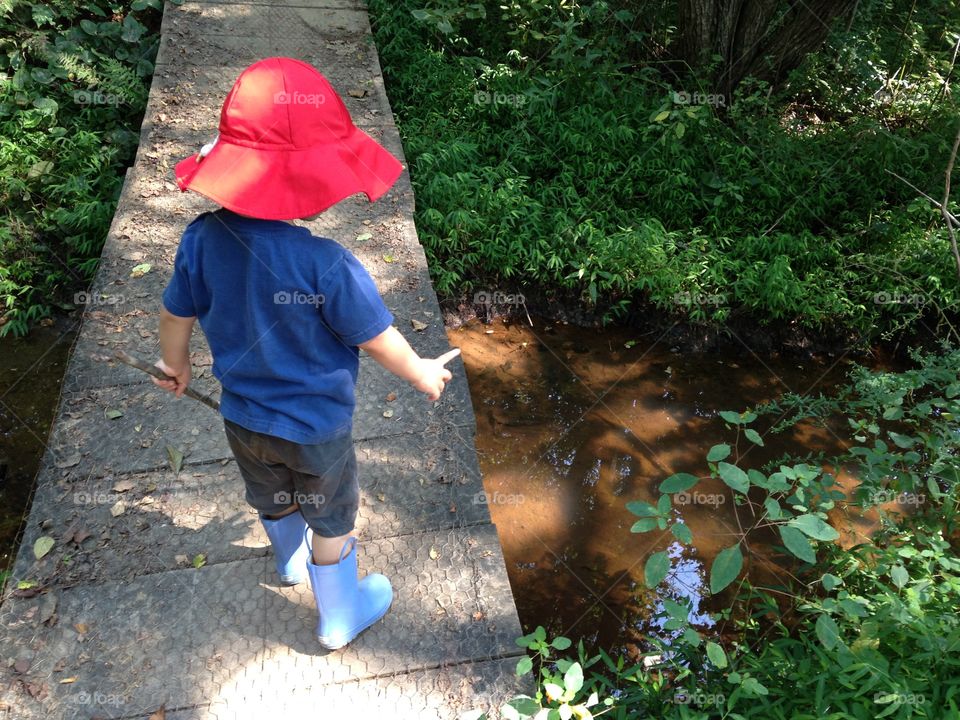 A two year old boy standing on a foot bridge pointing to the creek below, holding a stick and wearing a red hat and blue rain boots.