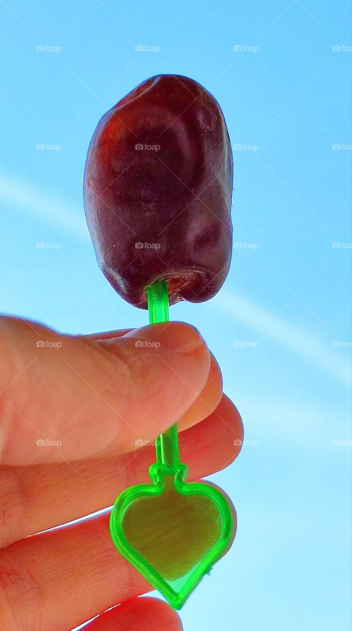 a woman's hand holds a green skewer, on which a purple date fruit. The contrast is created by a purple date fruit against a blue sky with white stripes from airplanes.