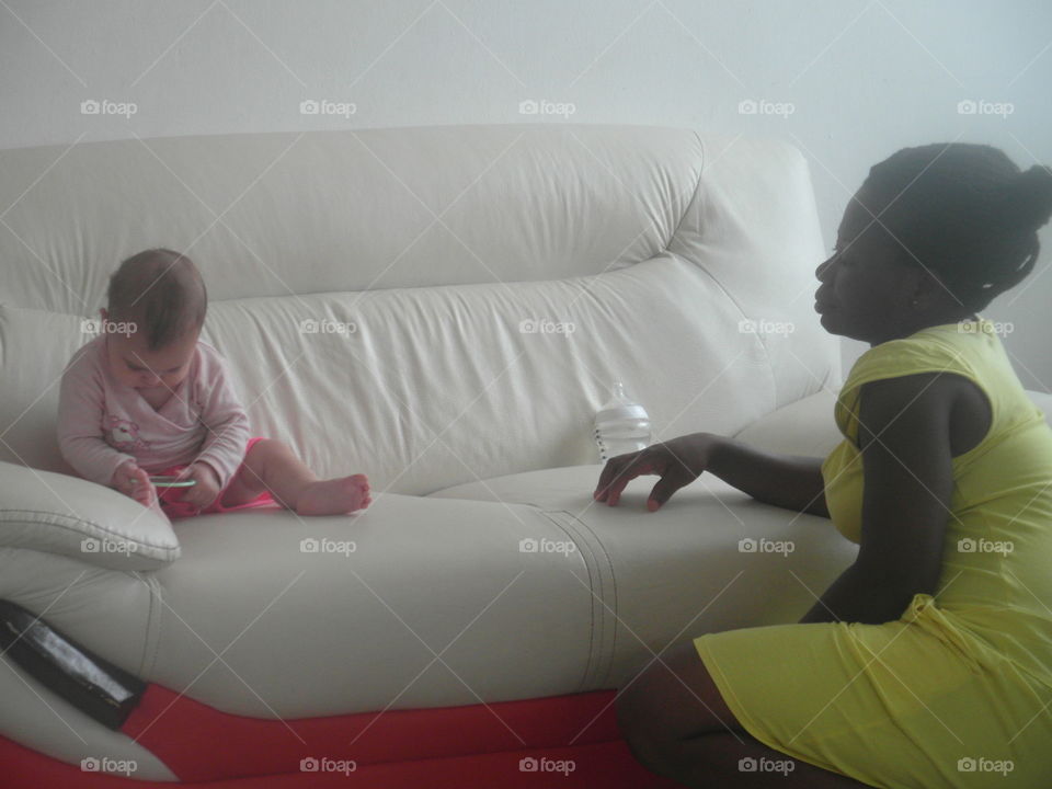 baby and Nanny. baby girl on leather couch exploring mirror while looked after by African Nanny