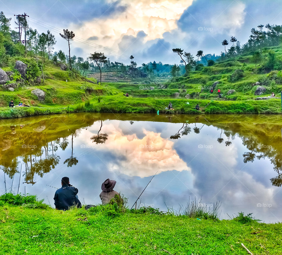 two people fishing at a lske.. reflection of clouds and green trees seen on the lake water