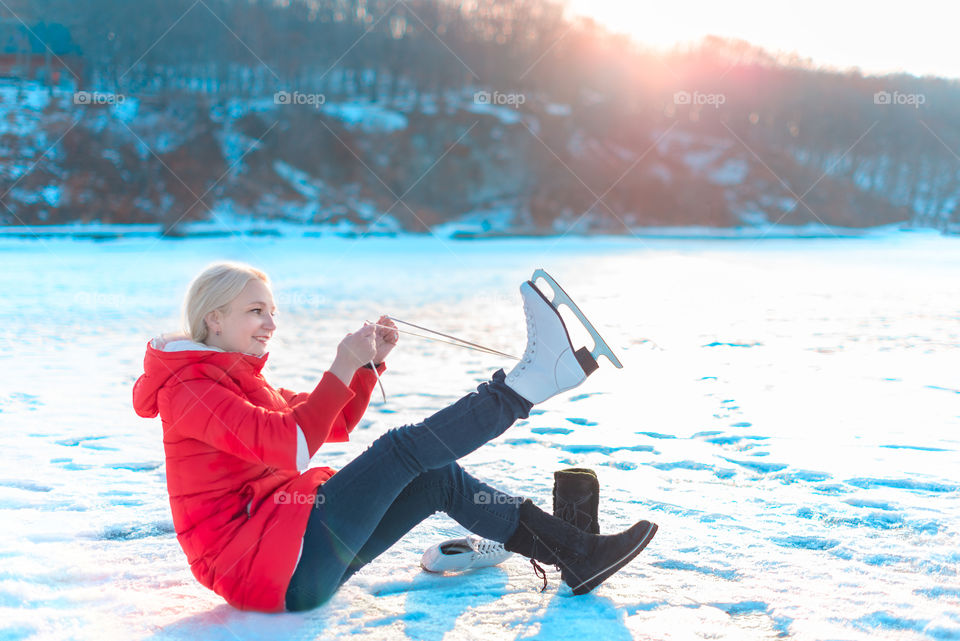 Young beautiful woman in red coat and figure skates sits on the frozen sea early in the morning. Winter outdoor activities concept. Leisure and lifestyle