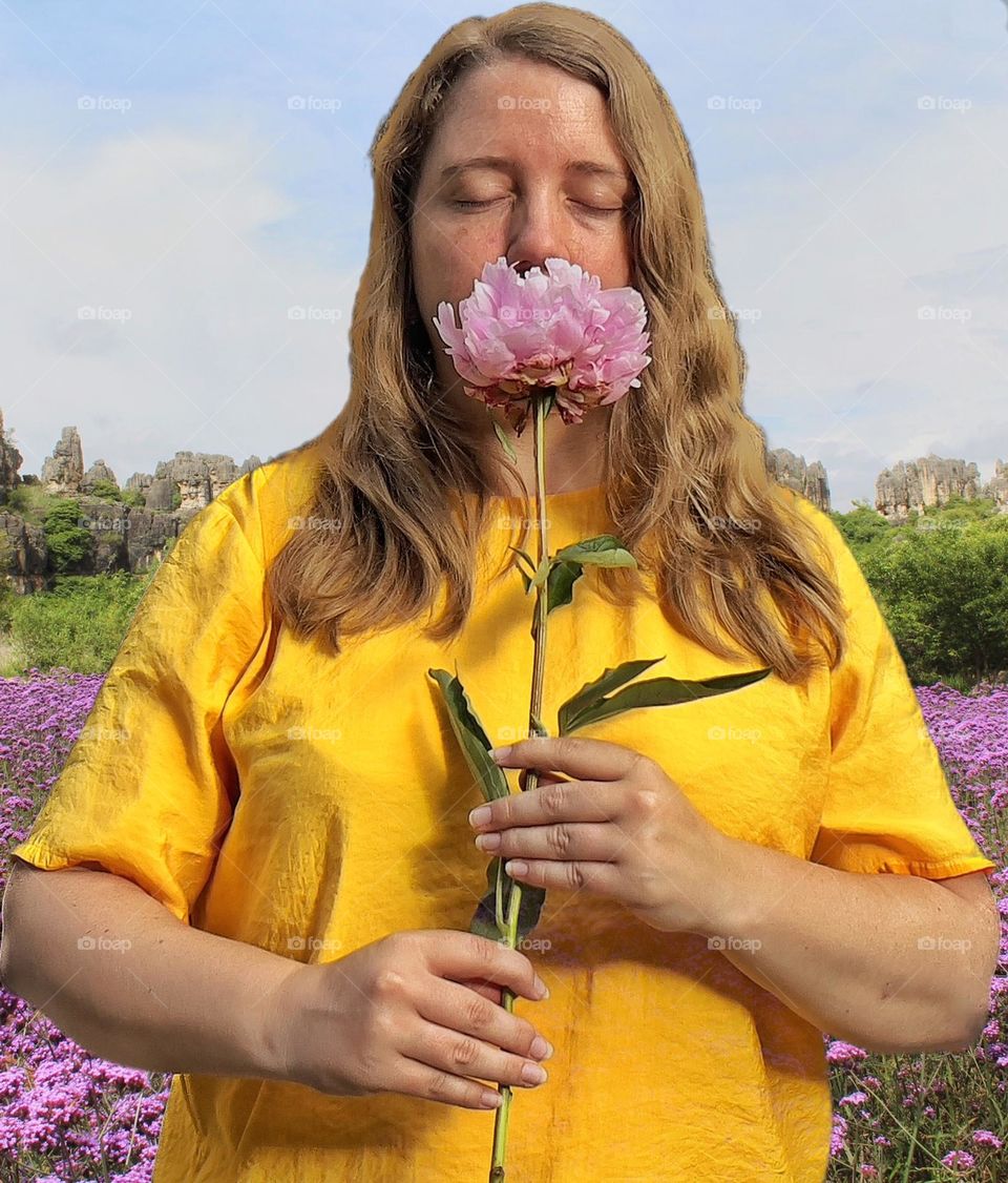 Double exposure of a woman smelling a peony while standing in a flower field with a stone forest in the background.