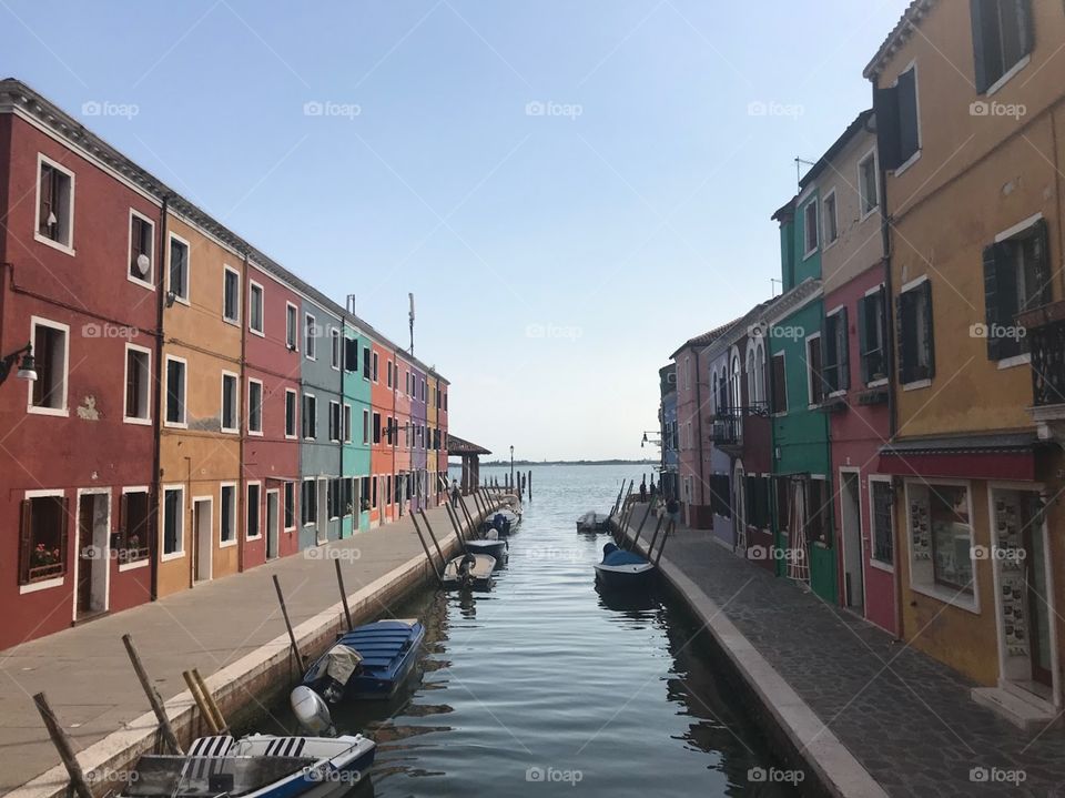 A small canal with a few canoes anchored, 2 sides are colorful houses, looking great. a pale blue sky with no clouds, no sunshine