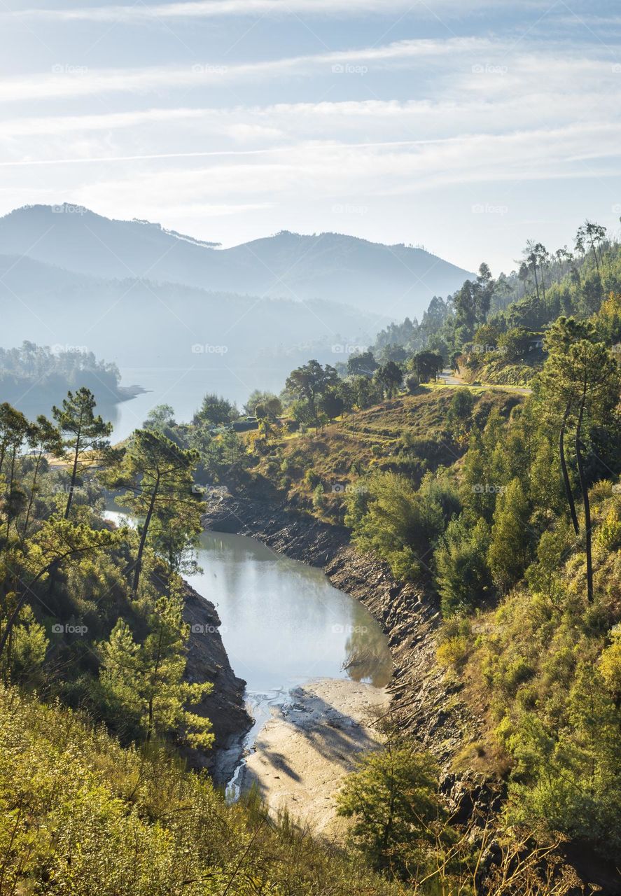 A river inlet leads through the tree covered valley, out to misty mountains in the distance 