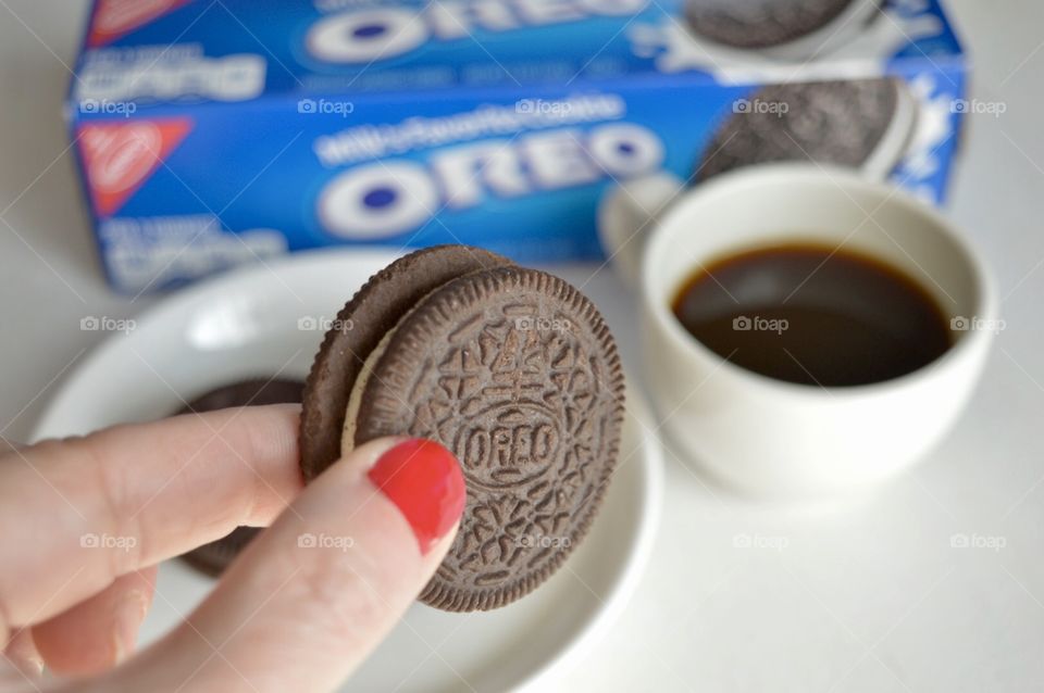 A woman’s fingers holding an Oreo cookie with a cup of coffee on a white background