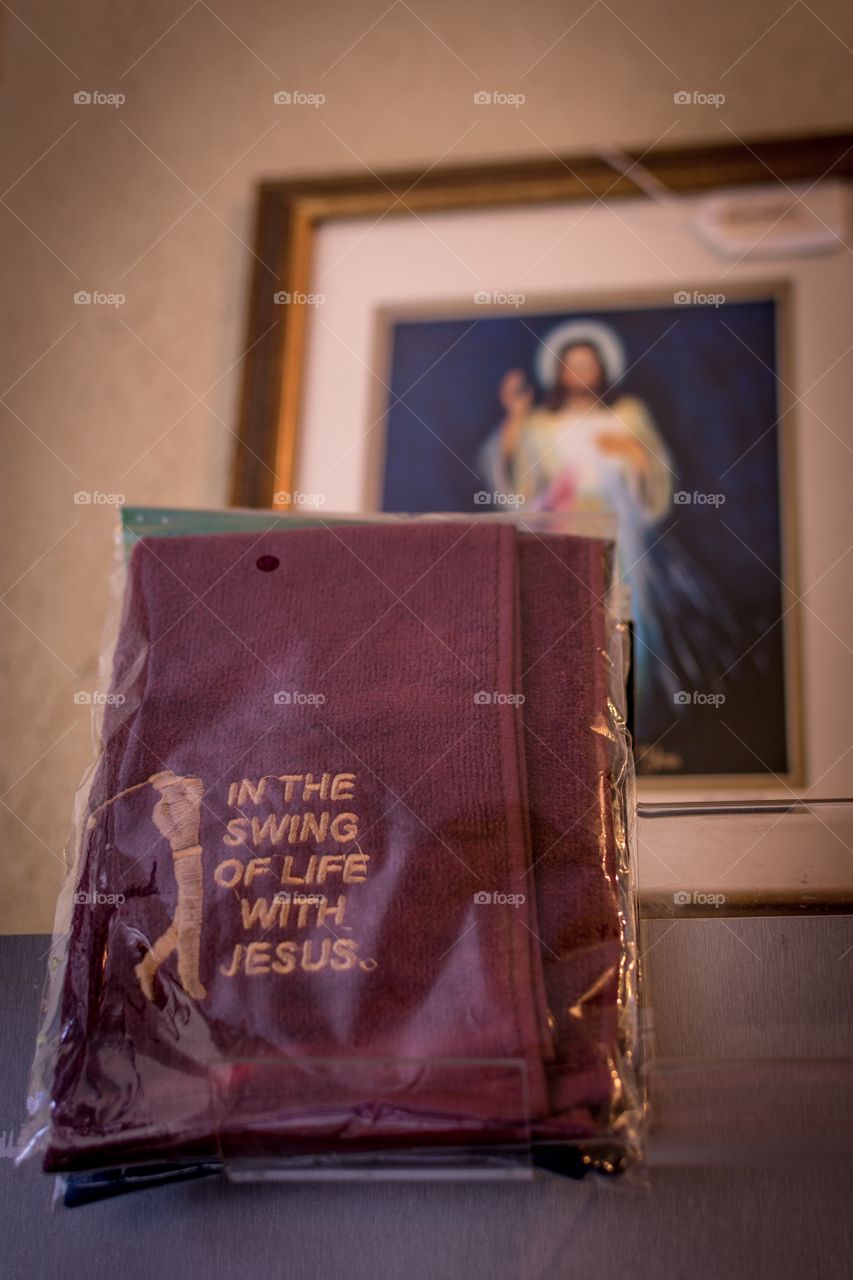 Plastic wrapped punny towel that is embroidered with the phrase “in the swing of life with Jesus” next to a golfer. Blurry Jesus painting overlooks it in the background.