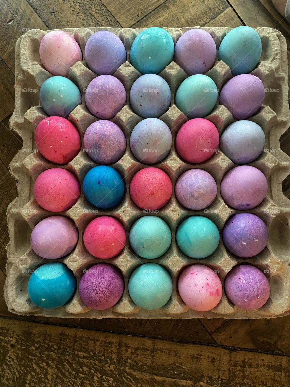 A variety of pastel, dye-colored Easter Eggs drying on an egg carton on the refrigerator shelf. The eggs are pink, purple, green, turquoise, blue, and teal. 