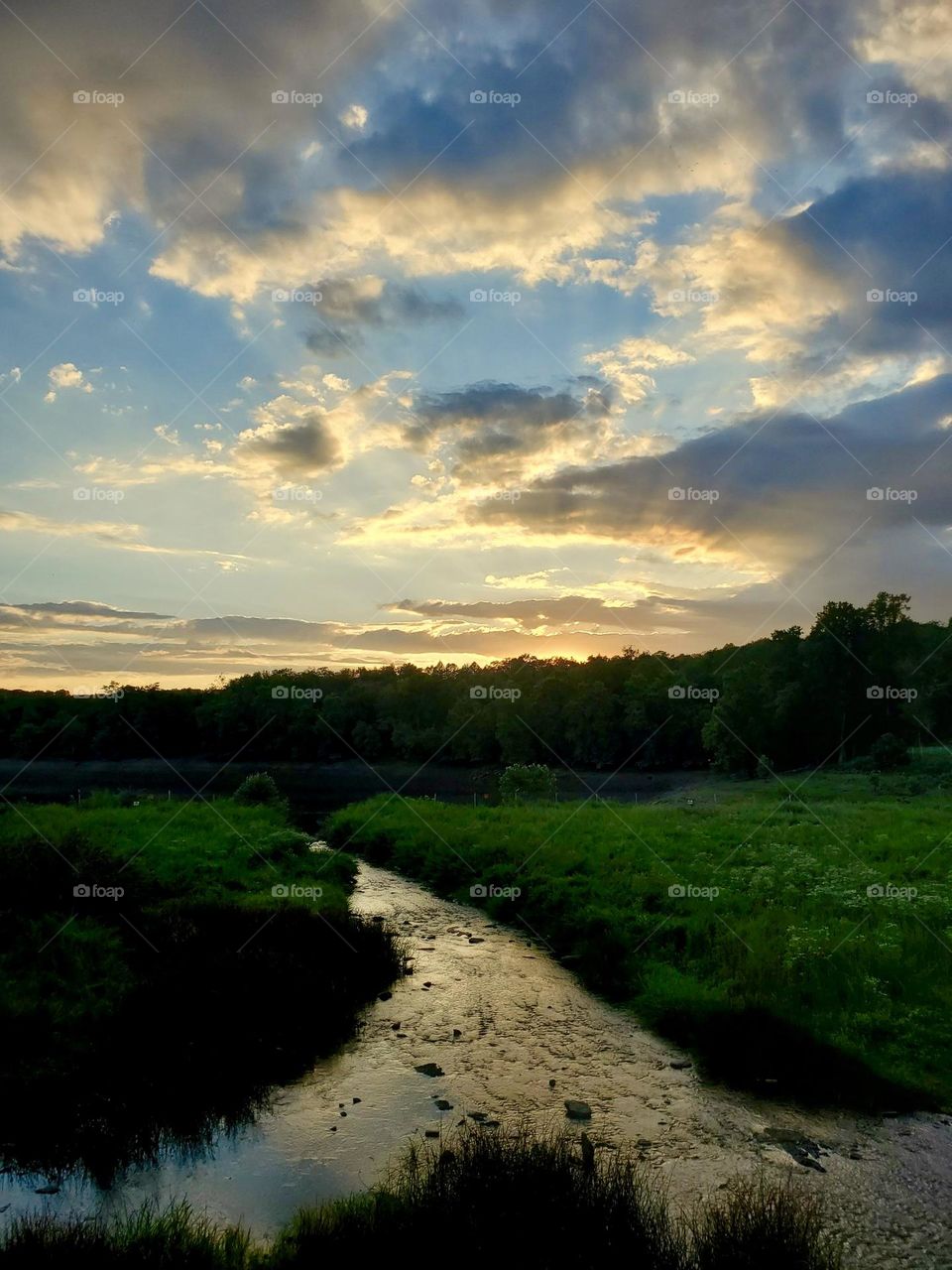 A gentle stream meandering along as the sun sets below the horizon, sending beams of light through the clouds. 