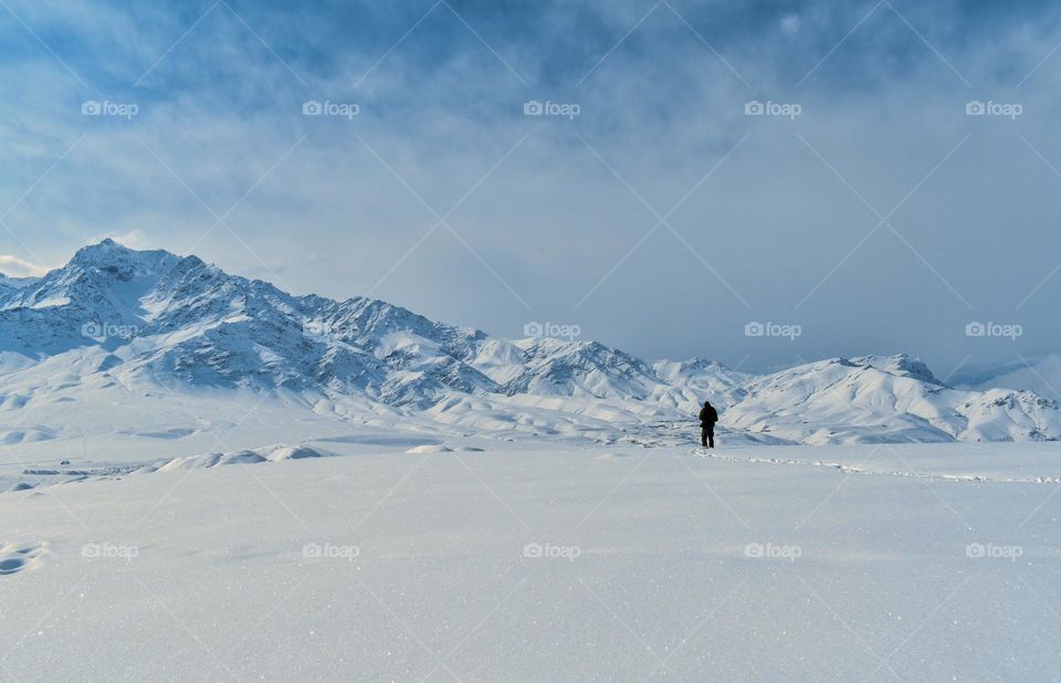 man walking on snow ;snow capped mountain ranges and blue sky in background .