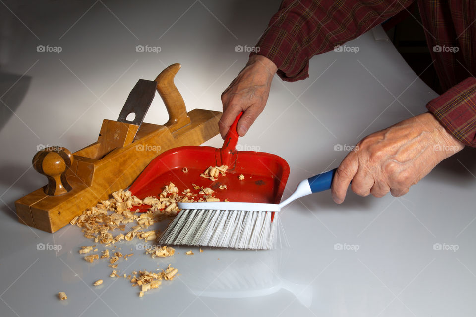 Hands of an old man clean up wooden chips from white surface with brush and dustpan after working with wooden planer (joiner).