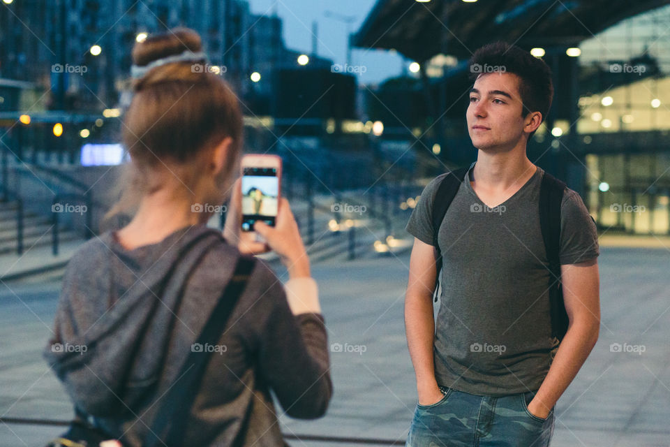 Young woman taking photos her friend, using a smartphone in the city at night