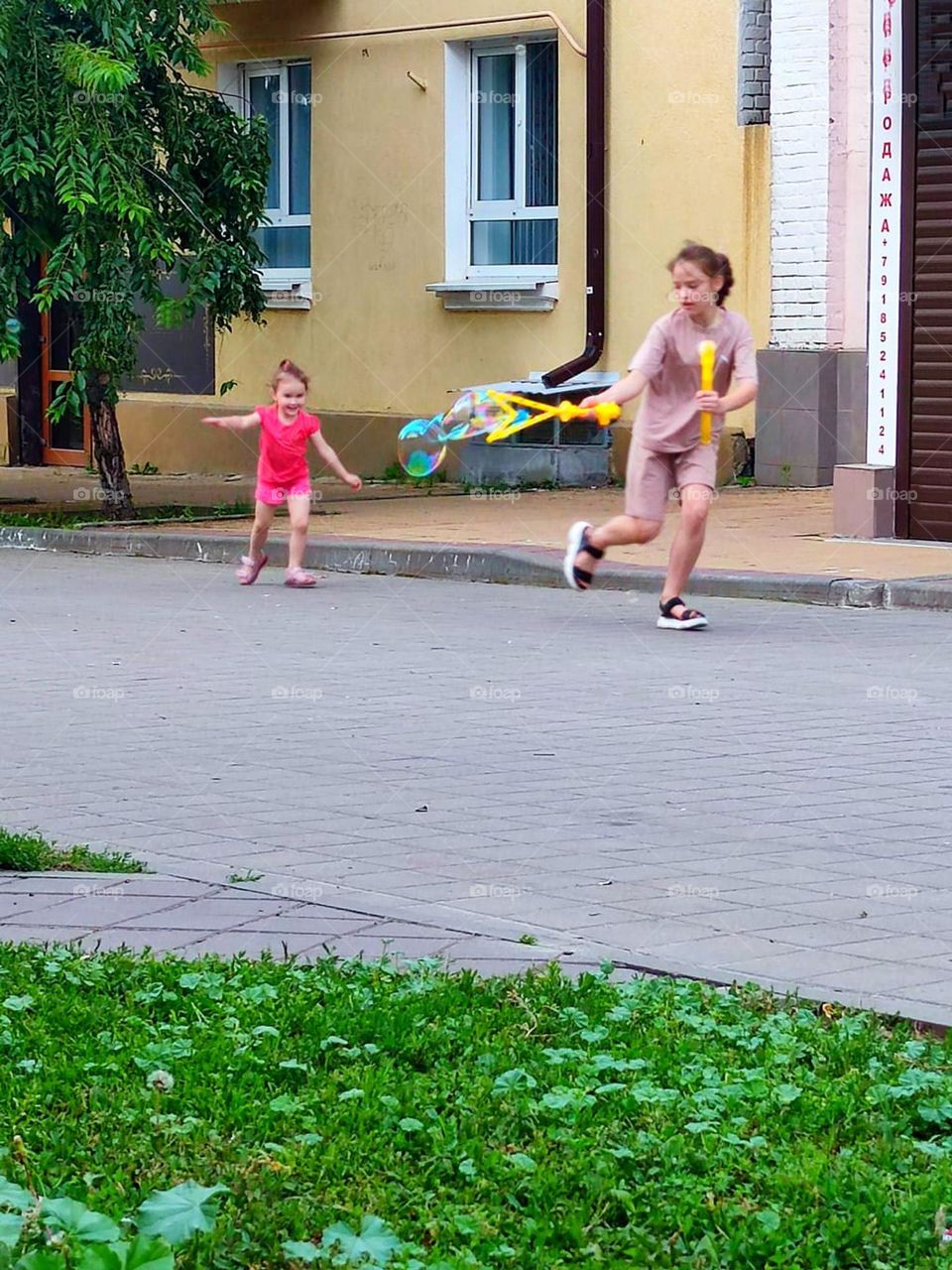 Girls run down the street.  In the hands of an older girl is a frame for soap bubbles.  A little girl runs after soap bubbles