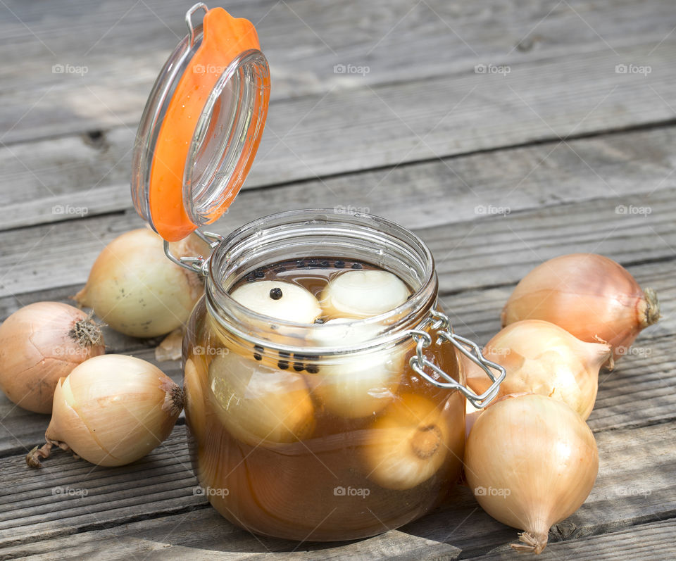 A jar of homemade pickled onions with the lid open on a textured wooden table surrounded by small unpeeled onions.