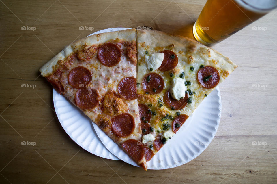 two slices of pizza with pepperoni ricotta and pesto on white plates with a beer