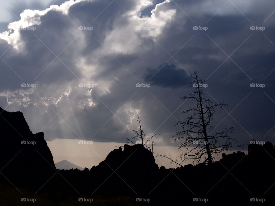 Beautiful sun rays break through the clouds after an evening summer rain storm at Smith Rocks in Central Oregon with Black Butte in the background and silhouetted trees in the foreground. 