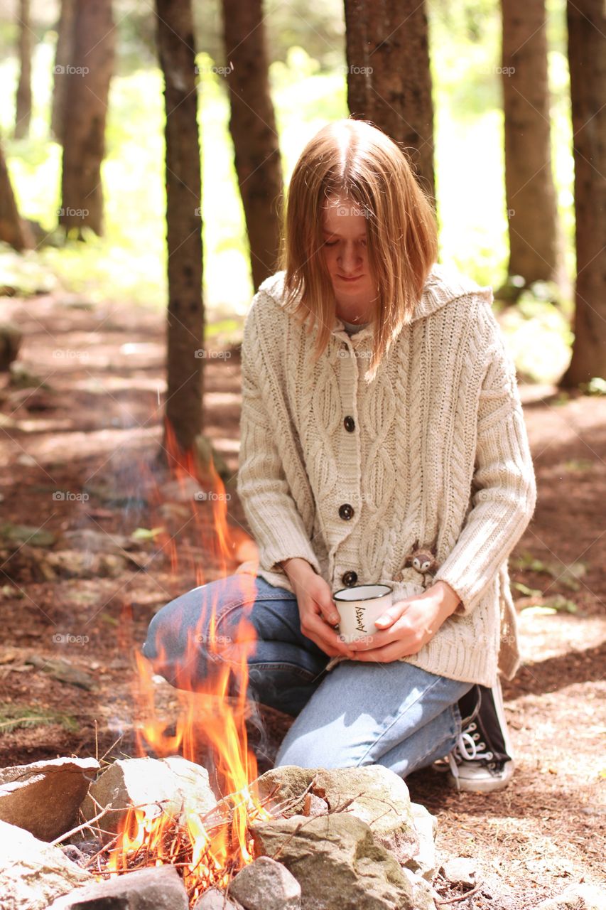 A girl with a cup of tea in her hands in a retinue sits by the fire in the mountains