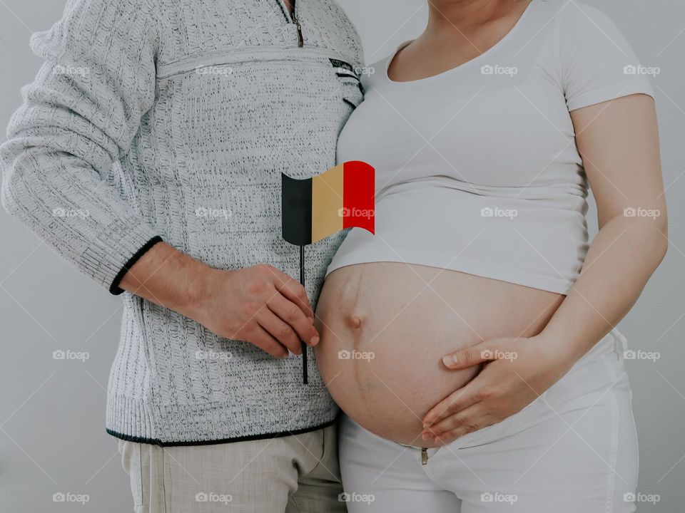 One young caucasian man and a pregnant woman with a bare belly stand in an embrace and hold a small paper Belgian flag in their hands, close-up side view.